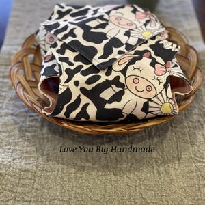 May include: Two fabric cow-print pouches with pink bows and daisy designs. The pouches are folded and placed in a woven basket. The text "Love You Big Handmade" is printed below the basket.