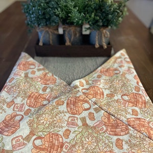 May include: A white fabric table runner with a repeating pattern of brown baskets filled with bread loaves, small jars, and white daisies. The text "Love You Big Handmade" is printed at the bottom of the runner.