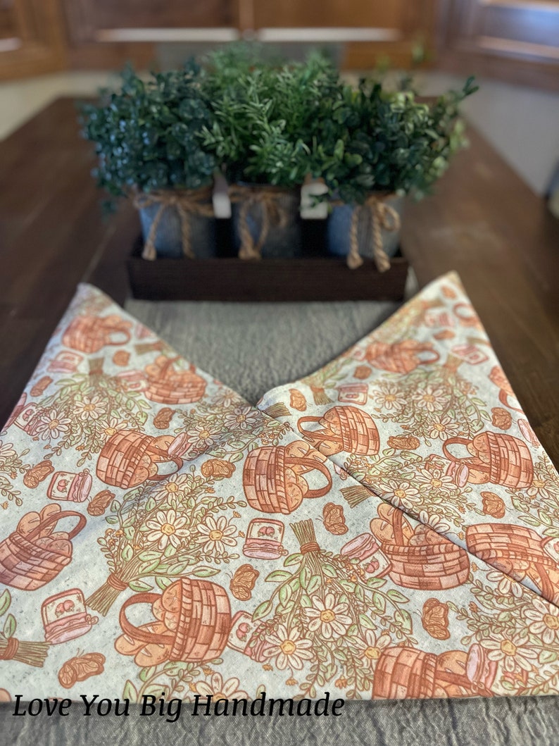 May include: A white fabric table runner with a repeating pattern of brown baskets filled with bread loaves, small jars, and white daisies. The text "Love You Big Handmade" is printed at the bottom of the runner.