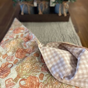 May include: A white and brown fabric table runner with a gingham pattern on the back. The front features a floral design with baskets of bread and jars of jam. The text "Love You Big Handmade" is printed on the bottom of the runner.