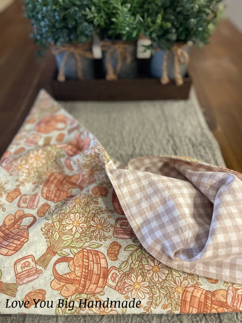 May include: A white and brown fabric table runner with a gingham pattern on the back. The front features a floral design with baskets of bread and jars of jam. The text "Love You Big Handmade" is printed on the bottom of the runner.