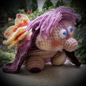 Könnte beinhalten: Ein gehäkeltes Stofftier mit einem rosa und violetten Körper, einer gelben und orangefarbenen Blume auf dem Rücken und blauen Augen. Das Tier sitzt auf einer hellbraunen Oberfläche.