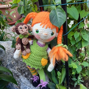 May include: A handmade crochet doll with orange hair in pigtails, wearing a green dress with yellow accents. The doll is holding a small brown crochet monkey. The doll's shoes are black with purple accents. The background features greenery.