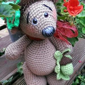 May include: A brown crocheted hedgehog toy with green and brown accents. The hedgehog is wearing a red ribbon and has large, black eyes. The toy is sitting on a wooden surface with green foliage in the background.