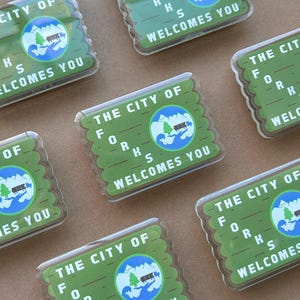 May include: Multiple rectangular green and clear plastic magnets. Each magnet has the text "THE CITY OF FORKS WELCOMES YOU" and a circular graphic with a tree, mountains, and water.