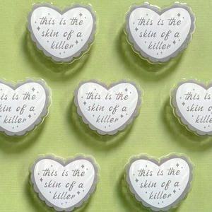 Peut inclure: Autocollants en forme de cœur avec un bord festonné et le texte "this is the skin of a killer" dans une police manuscrite. Les autocollants sont blancs avec des bordures et du texte gris, et sont éparpillés sur un fond vert clair.