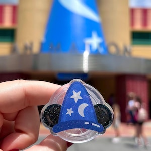 May include: A circular phone grip featuring a blue wizard hat design with white stars and a crescent moon, flanked by two black glittery mouse ears. The accessory has a clear plastic base and a glossy finish, showcasing a celestial theme.