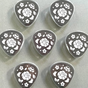 Pode incluir: Sete palhetas de guitarra pretas com um design floral branco. Cada palheta apresenta uma flor central rodeada por folhas e pequenos detalhes florais. As palhetas estão dispostas sobre uma superfície cinza claro.
