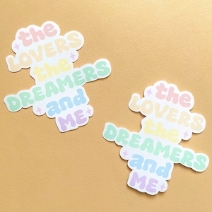 May include: Two pastel colored stickers with the text "the lovers the dreamers and me" in a rainbow gradient. The stickers are on a light brown background.