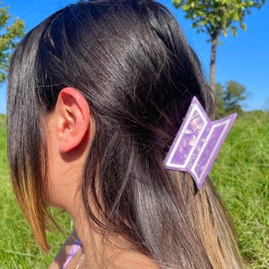 Puede incluir: Una pinza para el pelo morada y blanca está sujeta a un cabello castaño oscuro. La pinza tiene un diseño geométrico con un borde blanco. El fondo muestra hierba verde y un cielo azul.