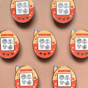 May include: A collection of orange and yellow enamel pins with a retro handheld game design. Each pin features the word "AVATAR" and symbols representing the elements of water, earth, air, and fire. The pins have a heart and star design.