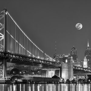 Peut inclure: Photographie en noir et blanc du pont Benjamin Franklin à Philadelphie la nuit. Le pont est illuminé, avec la ligne d'horizon de la ville en arrière-plan. Une pleine lune est visible dans le ciel nocturne au-dessus de la ville.