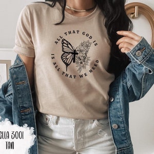 May include: A tan t-shirt with a black butterfly graphic and the text "All that God is is all that we need".