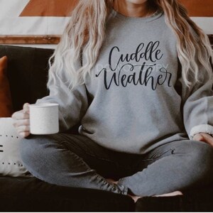 May include: A gray sweatshirt with the words "Cuddle Weather" in black script. The person is holding a white mug. The person is wearing gray jeans and sitting on a black couch with pillows.