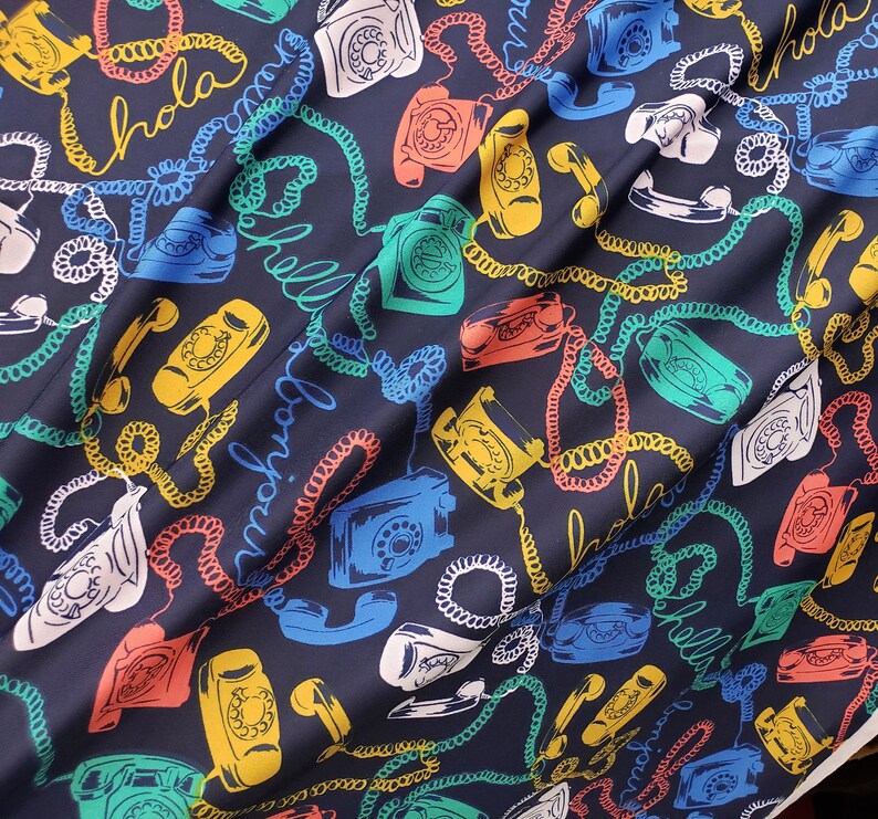 May include: A dark blue fabric with a repeating pattern of colorful rotary phones and coiled cords. The fabric has the word "Hola" written in a white cursive font.