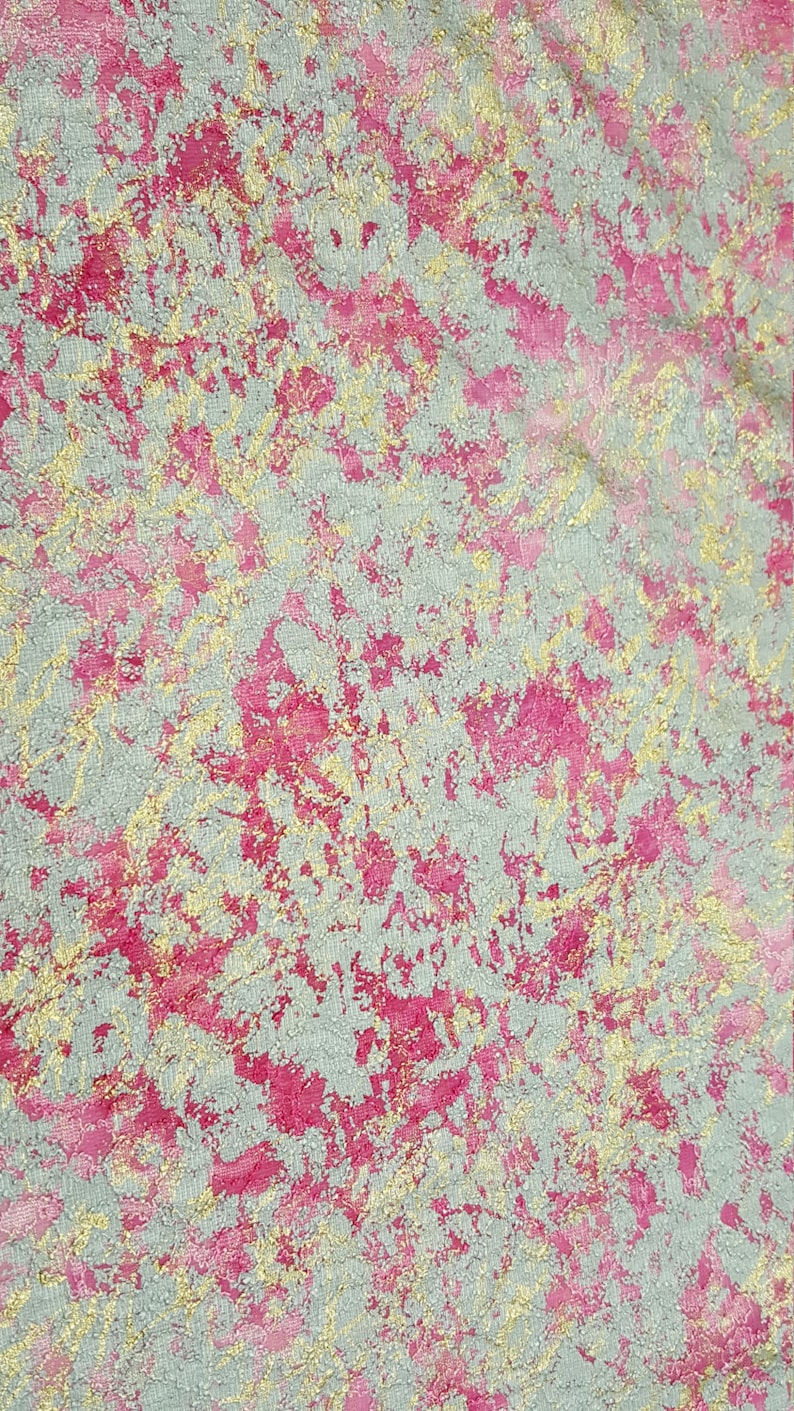May include: A close-up of a fabric with a textured surface. The fabric is a light gray color with pink and gold accents.