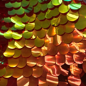 Pode incluir: Um close-up de um tecido vermelho coberto com lantejoulas douradas e verdes iridescentes dispostas em um padrão de escamas de peixe.