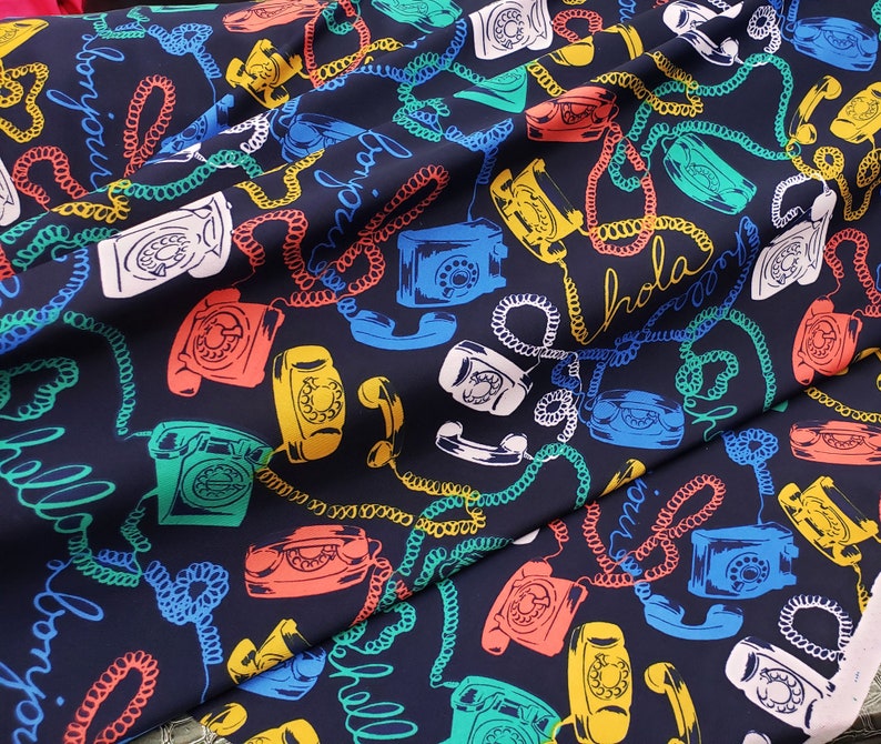 May include: A dark blue fabric with a repeating pattern of rotary phones and coiled cords. The phones are in various colors, including red, green, yellow, and white. The fabric has the words "Hola" and "Mama" printed on it.
