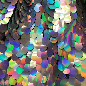 May include: Close-up of iridescent oval sequins in a variety of colors, arranged in a scaly pattern. The sequins have a holographic finish, creating a shimmering effect.