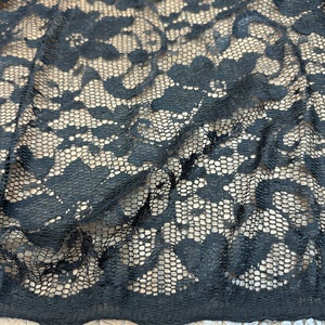 May include: Close-up of black lace fabric with a floral pattern. The lace is layered over a tan fabric, creating a contrast in color and texture. The intricate design of the lace is visible, showcasing the delicate details of the fabric.