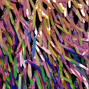 Puede incluir: Primer plano de lentejuelas iridiscentes en una variedad de colores, incluyendo morado, verde, rosa y amarillo. Las lentejuelas están dispuestas en un patrón aleatorio y están unidas a una tela negra.