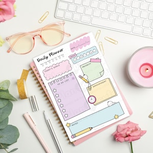 Puede incluir: Un planificador diario con espiral y cubierta rosa, con secciones para objetivos, listas de tareas y citas. La imagen incluye un teclado, gafas, bol&iacute;grafos, una vela y flores, creando una est&eacute;tica de espacio de trabajo.