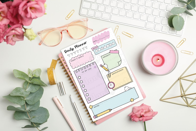 Puede incluir: Un planificador diario con espiral y cubierta rosa, con secciones para objetivos, listas de tareas y citas. La imagen incluye un teclado, gafas, bol&iacute;grafos, una vela y flores, creando una est&eacute;tica de espacio de trabajo.