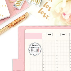May include: A pink planner with a gray and white December calendar sticker. The calendar sticker has a cloud-like shape with a scalloped edge. The planner has a grid pattern and is divided into two sections labeled "MON" and "TUES".