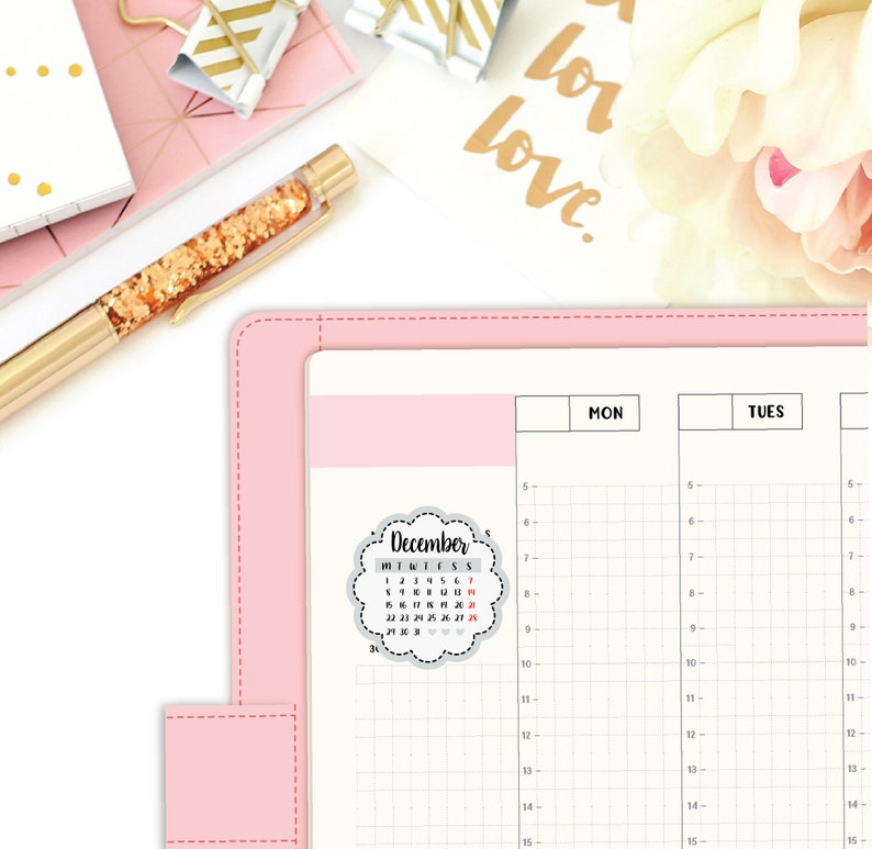 May include: A pink planner with a gray and white December calendar sticker. The calendar sticker has a cloud-like shape with a scalloped edge. The planner has a grid pattern and is divided into two sections labeled "MON" and "TUES".