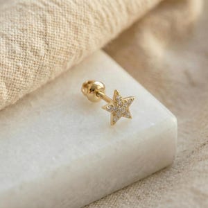 May include: A gold-colored star-shaped earring with small, clear gemstones. The earring is on a white surface, with a beige fabric in the background. The earring has a screw-back closure.