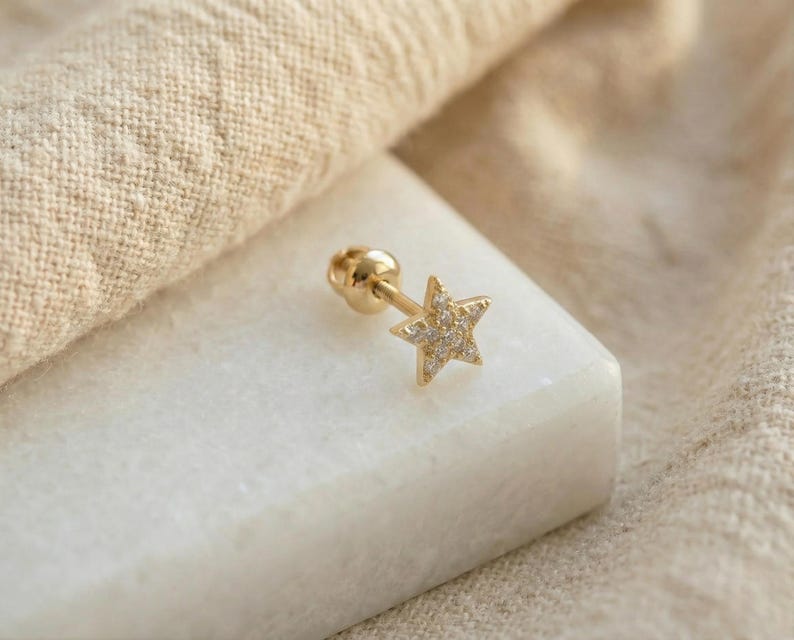 May include: A gold-colored star-shaped earring with small, clear gemstones. The earring is on a white surface, with a beige fabric in the background. The earring has a screw-back closure.