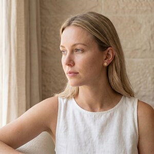 Puede incluir: Una mujer rubia con una camiseta sin mangas blanca y peque&ntilde;os pendientes de oro. El fondo muestra una ventana con cortinas transparentes y una pared de piedra. Los pendientes son de dise&ntilde;o sencillo y circular.
