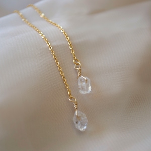 以下が含まれることがあります： 生のクリアクォーツクリスタルのドロップが付いた、繊細なゴールドチェーンのイヤリング。イヤリングは垂直に吊り下げられ、クリスタルの自然な美しさを際立たせています。背景は柔らかくニュートラルな生地で、ジュエリーのエレガントさを際立たせています。