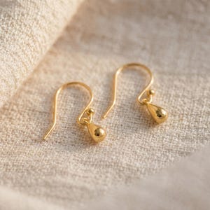 Può includere: Un paio di orecchini a goccia color oro. Ogni orecchino presenta un gancio curvo e una piccola perla rotonda sopra un ciondolo a forma di goccia. Gli orecchini sono esposti su un tessuto strutturato bianco sporco.