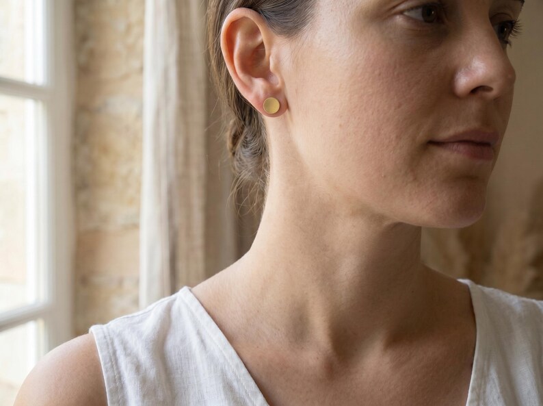 Puede incluir: Primer plano de una persona que lleva un pendiente de bot&oacute;n circular de color dorado. El pendiente tiene un dise&ntilde;o sencillo y minimalista. La persona lleva una camiseta blanca.