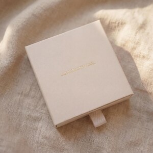 Puede incluir: Una caja cuadrada de color beige claro con una cinta para tirar, sobre una tela beige. La caja tiene letras doradas en la parte superior. La tela tiene un aspecto texturizado, con sombras y reflejos.