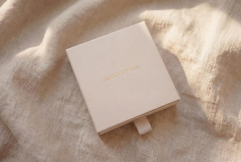 Puede incluir: Una caja cuadrada de color beige claro con una cinta para tirar, sobre una tela beige. La caja tiene letras doradas en la parte superior. La tela tiene un aspecto texturizado, con sombras y reflejos.