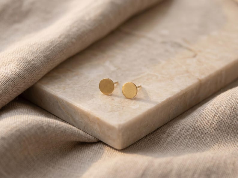 Puede incluir: Un par de pendientes de oro con dise&ntilde;o circular. Los pendientes se muestran sobre una superficie rectangular de color claro, con una tela de tono neutro en el fondo. Los pendientes son peque&ntilde;os y minimalistas.