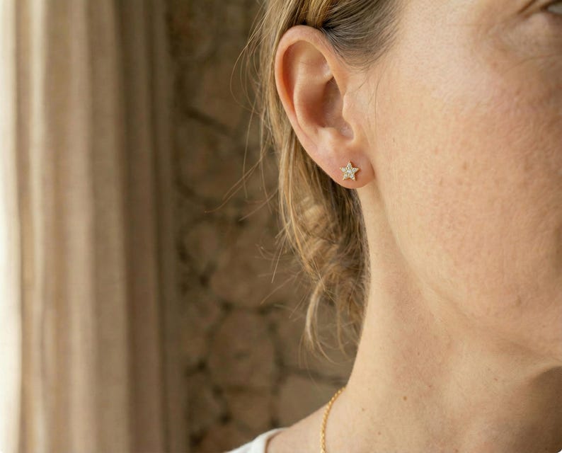 May include: Close-up of a person's ear adorned with a small, gold star-shaped earring. The earring is studded with tiny, sparkling gems. The person's skin is fair, and their hair is pulled back, revealing the ear and earring.