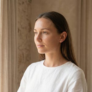May include: A woman wearing a white shirt with a star-shaped earring in her ear. The earring is gold-colored and small. The woman has long brown hair and is looking off to the side.