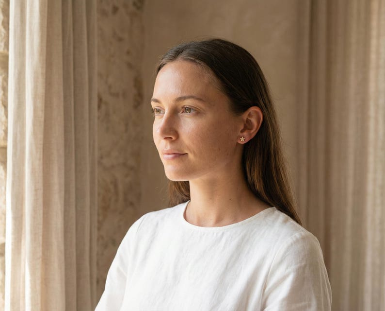 May include: A woman wearing a white shirt with a star-shaped earring in her ear. The earring is gold-colored and small. The woman has long brown hair and is looking off to the side.