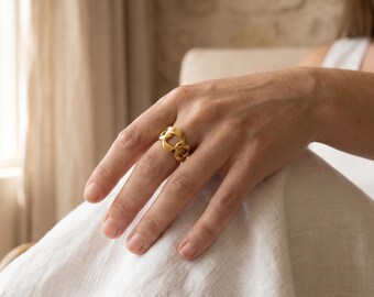 May include: A gold-colored ring with a chain link design. The ring is worn on a finger, and the hand is resting on a white fabric. The ring's links are interlocked, creating a bold and stylish look. The ring is a statement piece.