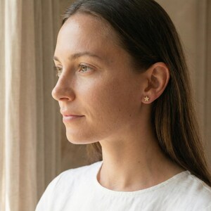 May include: A woman wearing a white shirt with a pair of gold star-shaped stud earrings. The earrings are small and delicate, adding a touch of sparkle to the ear. The woman is looking off to the side.