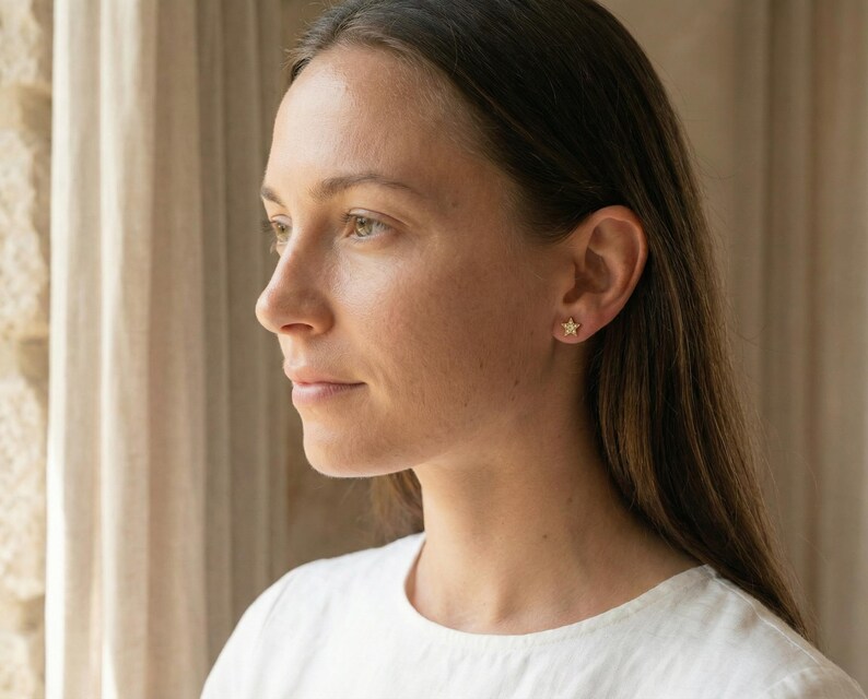 May include: A woman wearing a white shirt with a pair of gold star-shaped stud earrings. The earrings are small and delicate, adding a touch of sparkle to the ear. The woman is looking off to the side.