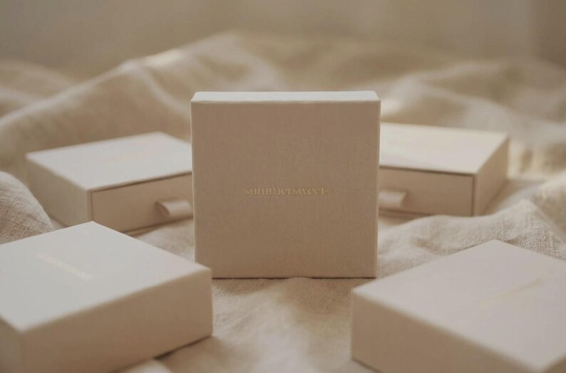 Puede incluir: Cuatro cajas rectangulares blanquecinas, una de pie, las otras planas. Las cajas est&aacute;n dispuestas sobre una superficie clara y texturizada. El texto "something sweet" est&aacute; en relieve en la parte delantera de la caja vertical. Dise&ntilde;o minimalista.