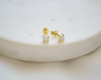 May include: A pair of small stud earrings with a gold setting and square, white opal stones. The earrings are displayed on a white, marble-like surface. The studs are a delicate jewelry item.
