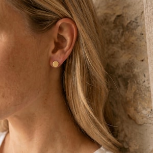 Puede incluir: Primer plano de la oreja de una mujer con un pendiente de bot&oacute;n dorado. El pendiente es un peque&ntilde;o disco redondo. La mujer tiene el pelo rubio y lleva una camisa blanca. El fondo es una pared texturizada y una cortina beige.