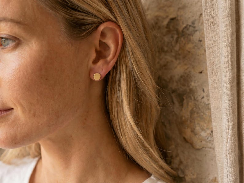 Puede incluir: Primer plano de la oreja de una mujer con un pendiente de bot&oacute;n dorado. El pendiente es un peque&ntilde;o disco redondo. La mujer tiene el pelo rubio y lleva una camisa blanca. El fondo es una pared texturizada y una cortina beige.