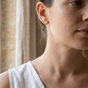 Puede incluir: Primer plano de una oreja con un pendiente de bot&oacute;n redondo dorado. El pendiente tiene un dise&ntilde;o simple y minimalista. La persona lleva una camiseta sin mangas blanca.