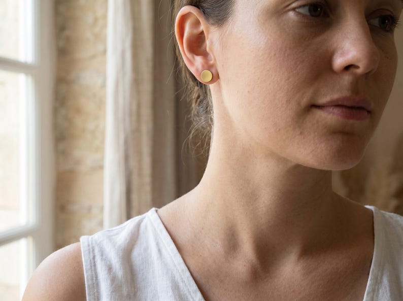 Puede incluir: Primer plano de una oreja con un pendiente de bot&oacute;n redondo dorado. El pendiente tiene un dise&ntilde;o simple y minimalista. La persona lleva una camiseta sin mangas blanca.